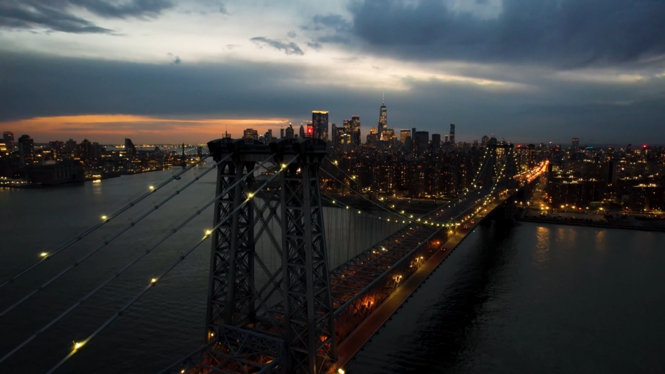 A steel suspension bridge at dusk, the Manhattan skyline behind it, seen from a drone